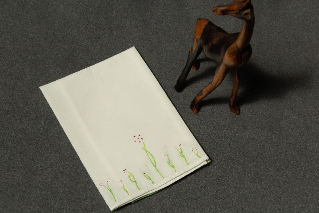 A folded handkerchief painted with small plants with plum colored flowers, next to a small wooden giraffe