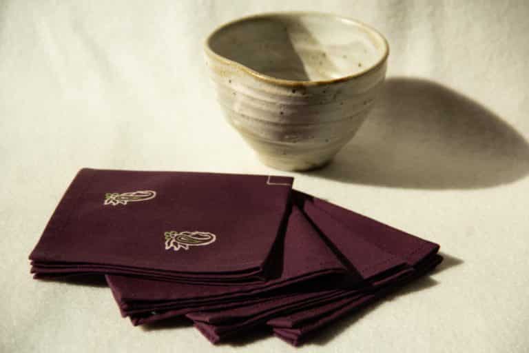 Four deep purple cocktail napkins with cream and green prints next to a ceramic cup.