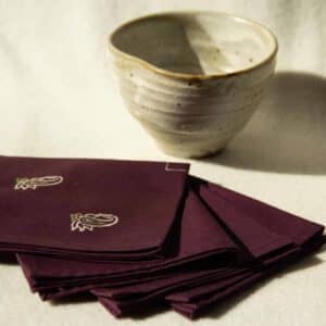 Four deep purple cocktail napkins with cream and green prints next to a ceramic cup.