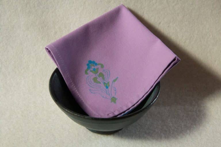 A lilac colored cocktail napkin with a blue and green floral print folded inside a bowl.