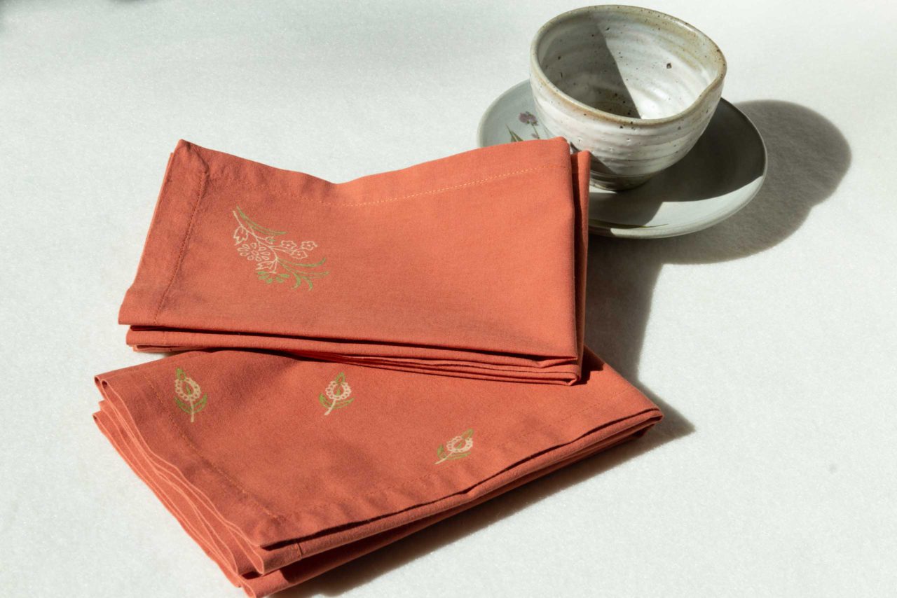 A set of napkins in terracotta with green and cream prints next to a ceramic cup and dish.