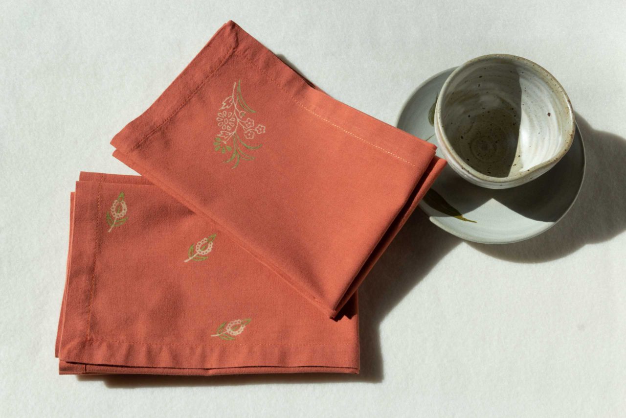 A set of napkins in terracotta color with green and cream prints next to a ceramic cup and plate