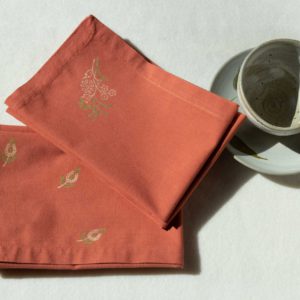 A set of napkins in terracotta color with green and cream prints next to a ceramic cup and plate